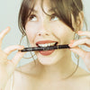 Une jeune femme brune de 25 ans, au teint clair, cadrée en gros plan, tient un stylo eyeliner adhésif 2 en 1 noir, le BlackFadime de la marque FADIME. Avec humour, elle le serre entre ses dents, regardant vers le haut et à droite. BlackFadime offre une pointe fine, feutre souple, haute précision, tenue parfaite. Waterproof et végan, il assure fixation rapide, tenue durable, grâce à ses polymères naturels. Un allié ultra précis