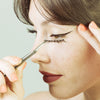 Jeune femme brune, 25 ans de profil gauche, au teint clair, elle tient la pince du kit d'application FADIME, faux cil en chanvre 100% végétal, biodégradable, bandes transparentes en coton. Émerveillée, amène la pince vers son œil. Avec tracé magnifique et précis BlackFadime, eyeliner adhésif 2 en 1 noir. Haute précision, tenue parfaite, Waterproof, végan, fixation rapide, tenue durable. Polymères doux et durables, haute qualité, hypoallergéniques. Flexibles, confort sans paraben. ultra précis