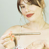 Une jeune femme brune de 25 ans, au teint clair, elle tient de la main droite le stylo eyeliner adhésif transparent et le présente de la main gauche le Clearfadime de FADIME avec un jolie sourire . Sa pointe fine et feutre souple garantit haute précision. Waterproof, végan, il assure tenue parfaite et rapide fixation grâce aux polymères naturels. Hypoallergénique. Un allié ultra précis pour un regard irrésistible.